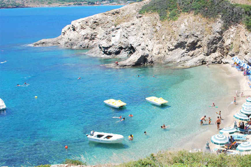 Clear shallow water and sandy shore at Bali Beach, Crete, with small fishing boats and rocky hills in the background