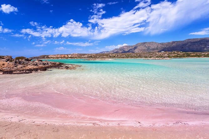 A wide view of Elafonisi Beach with shallow turquoise water, pale pink sand, and the lagoon area in early morning light