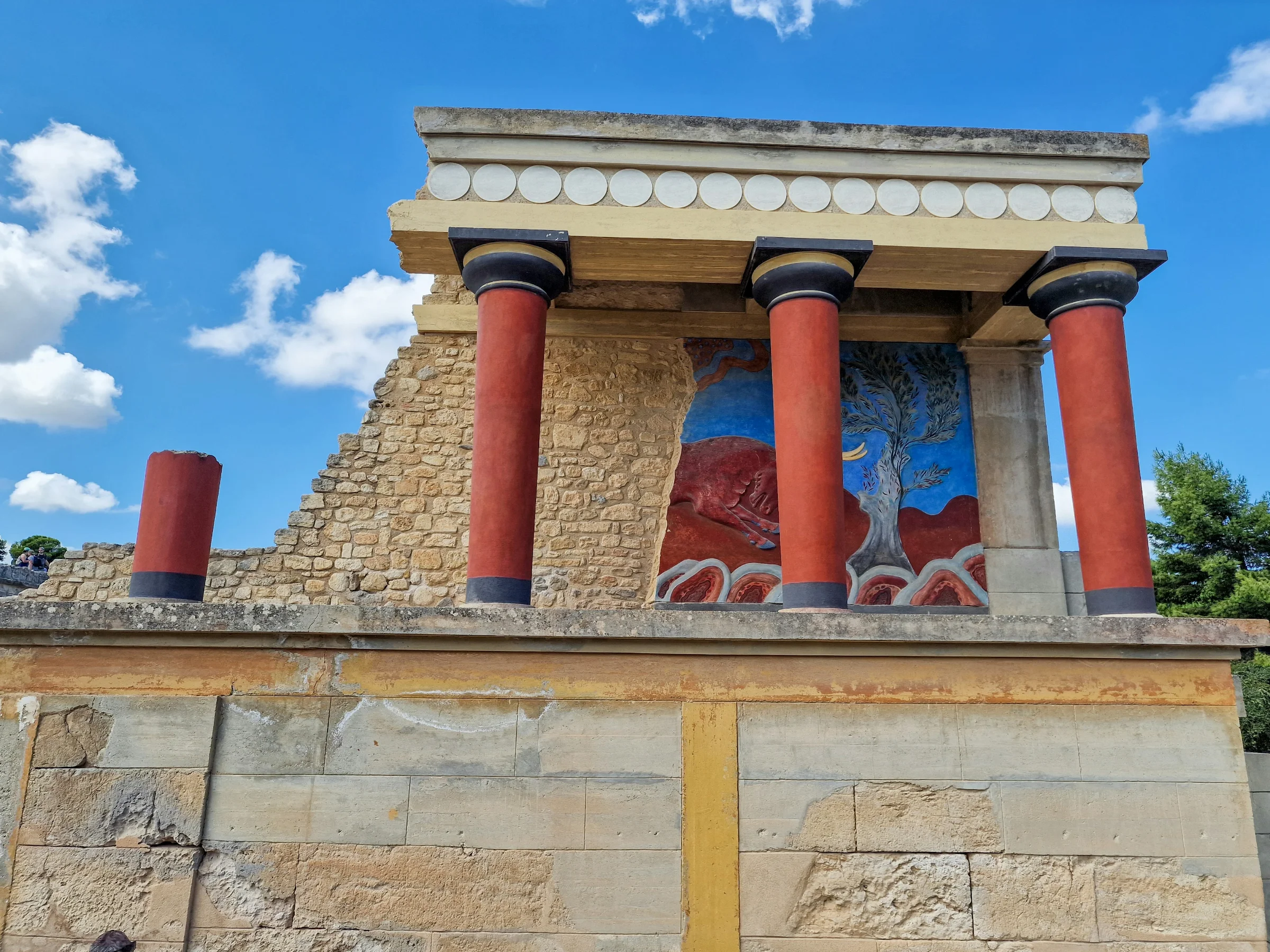 Wide view of Knossos Palace showing reconstructed red columns and the main palace complex under a clear sky, Heraklion, Crete